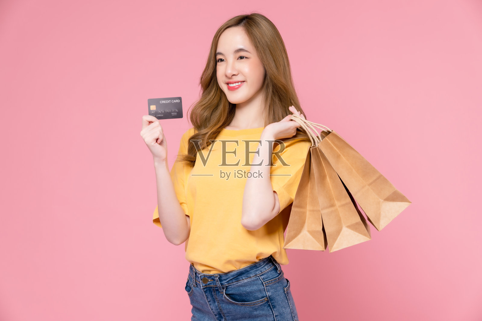 美丽的亚洲女人拿着棕色空白的牛皮纸购物袋和显示信用卡在粉红色的背景。照片摄影图片
