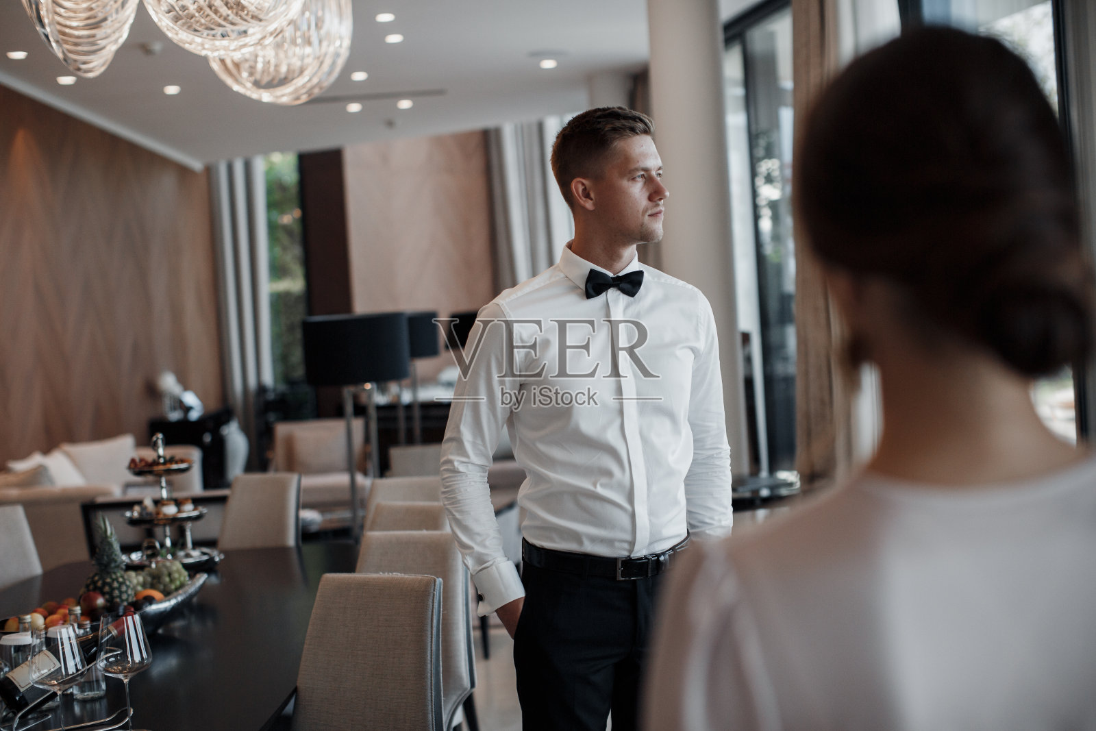 一个穿着白色礼服的美丽新娘和一个穿着白色衬衫的年轻新郎正在室内准备婚礼仪式。照片摄影图片