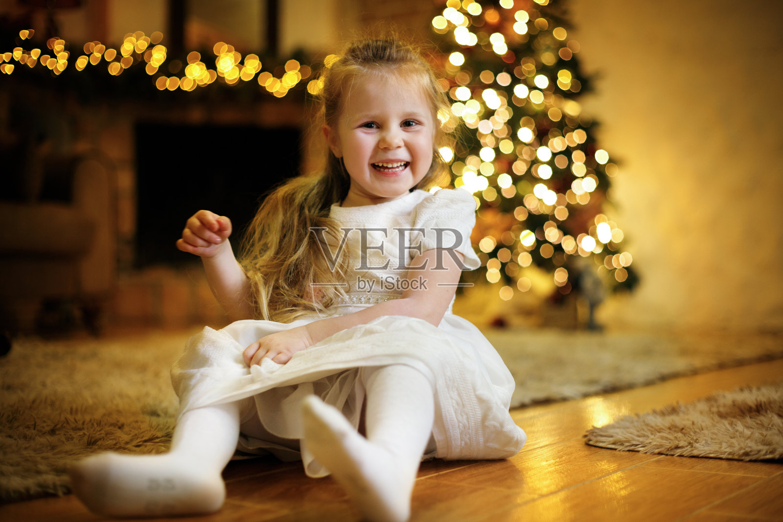 快乐可爱的女孩穿着白色的裙子在圣诞树的圣诞室内地板上摆姿势照片摄影图片