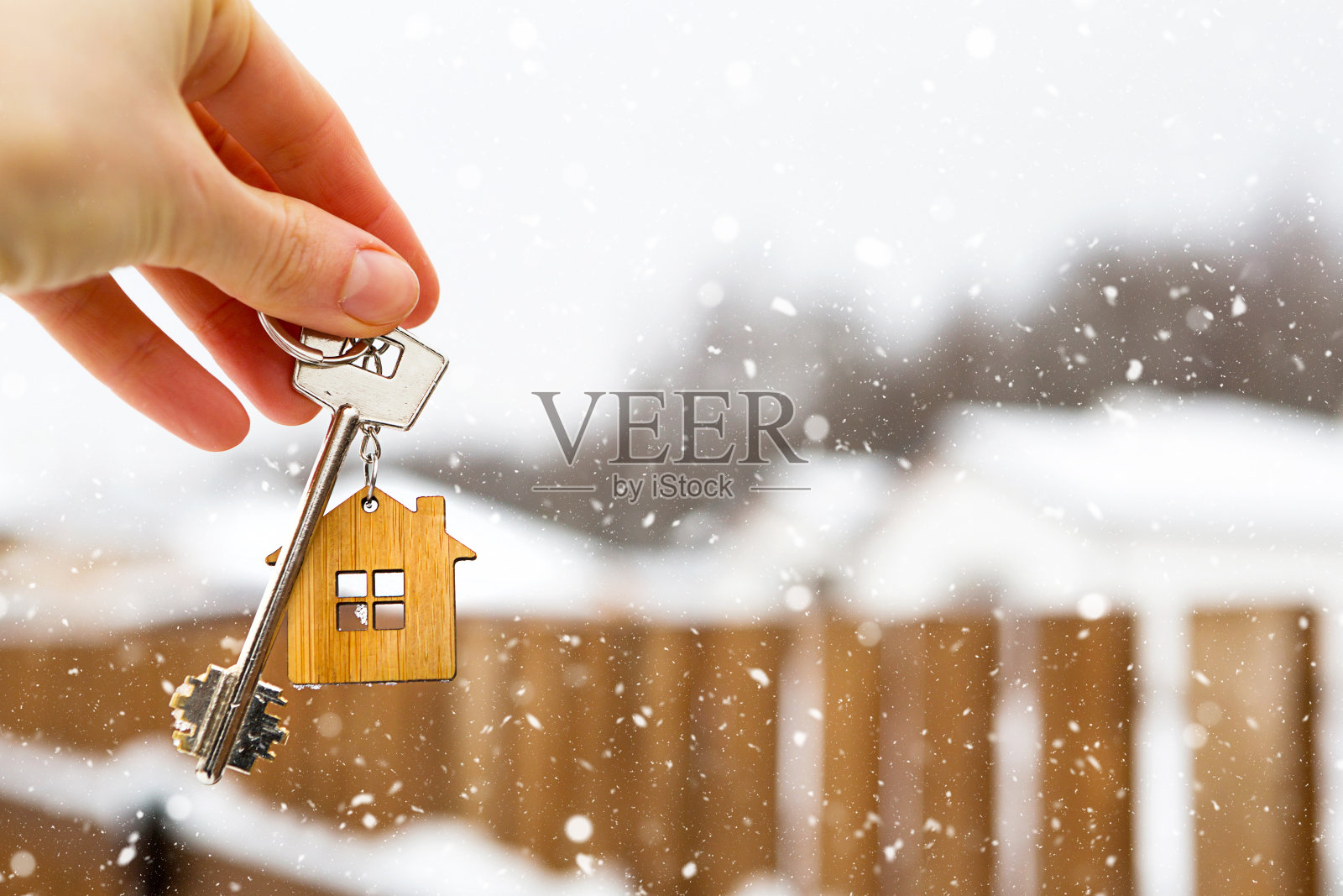 手拿钥匙的房子在冬天的雪地里。篱笆和小屋的背景。在新年和圣诞节送出一份礼物。搬到新房子，抵押，租赁和购买房地产。开门。照片摄影图片