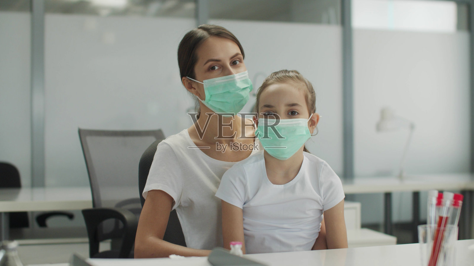 在白色的办公室里，一位母亲带着小女儿坐在椅子上，戴着医用口罩，对着镜头摆姿势照片摄影图片