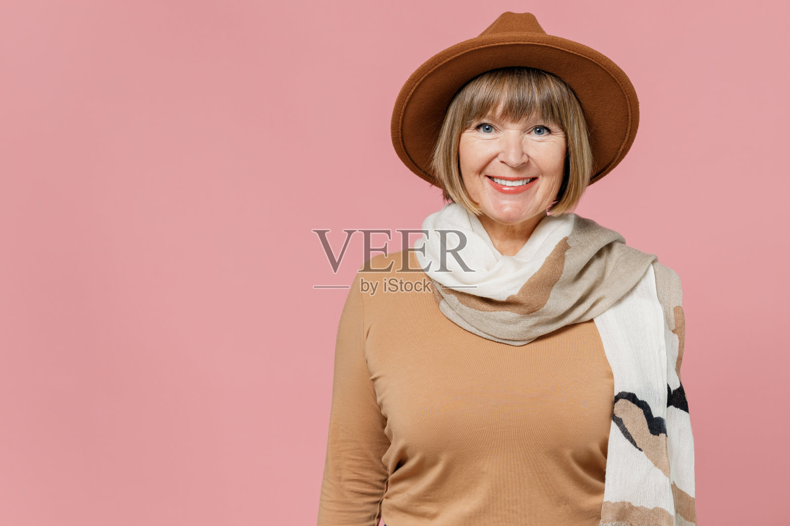 旅行者旅行者乐趣成熟的老年老年女士妇女55岁穿着棕色衬衫帽子围巾看相机孤立在素色淡粉色背景的摄影棚肖像。航空旅行概念照片摄影图片