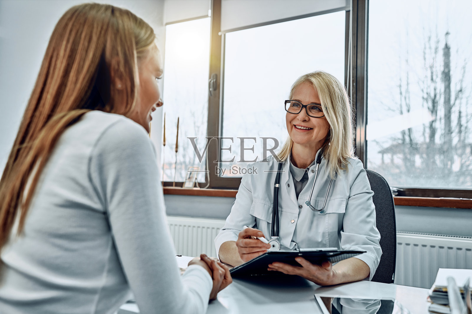 美丽的高级女医生和病人坐在工作桌前，在特殊表格上写处方。照片摄影图片