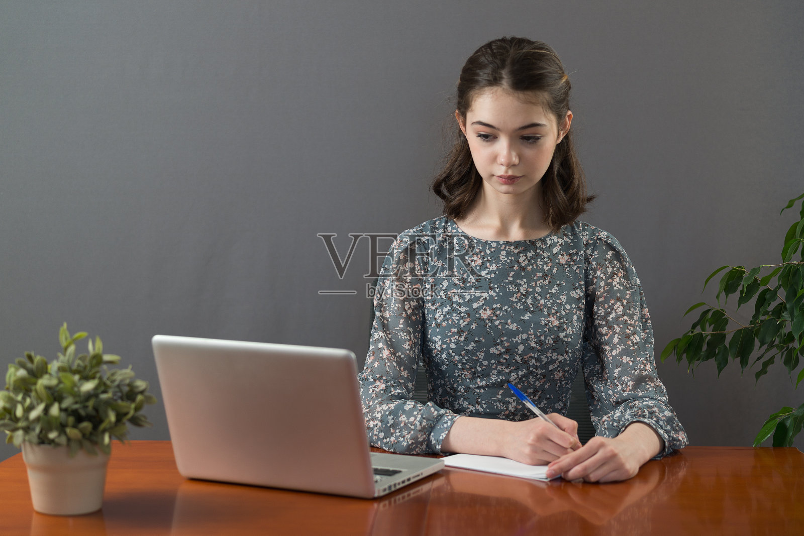 一位漂亮严肃的女学生，穿着灰色的裙子，坐在桌子旁，桌上放着笔记本电脑和用来记笔记的笔记本。独立备考照片摄影图片