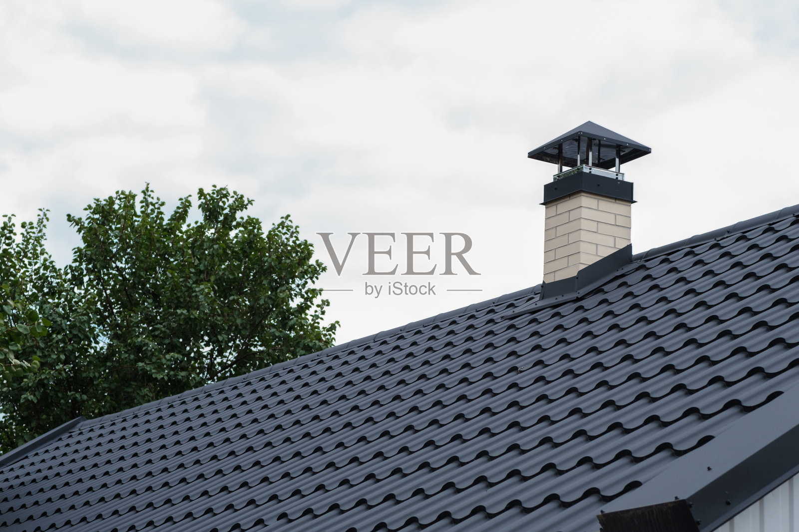 屋顶有深色的金属瓦和烟囱，金属瓦屋顶。黑色的金属屋顶上有一个烟囱，背景是多云的天空。照片摄影图片