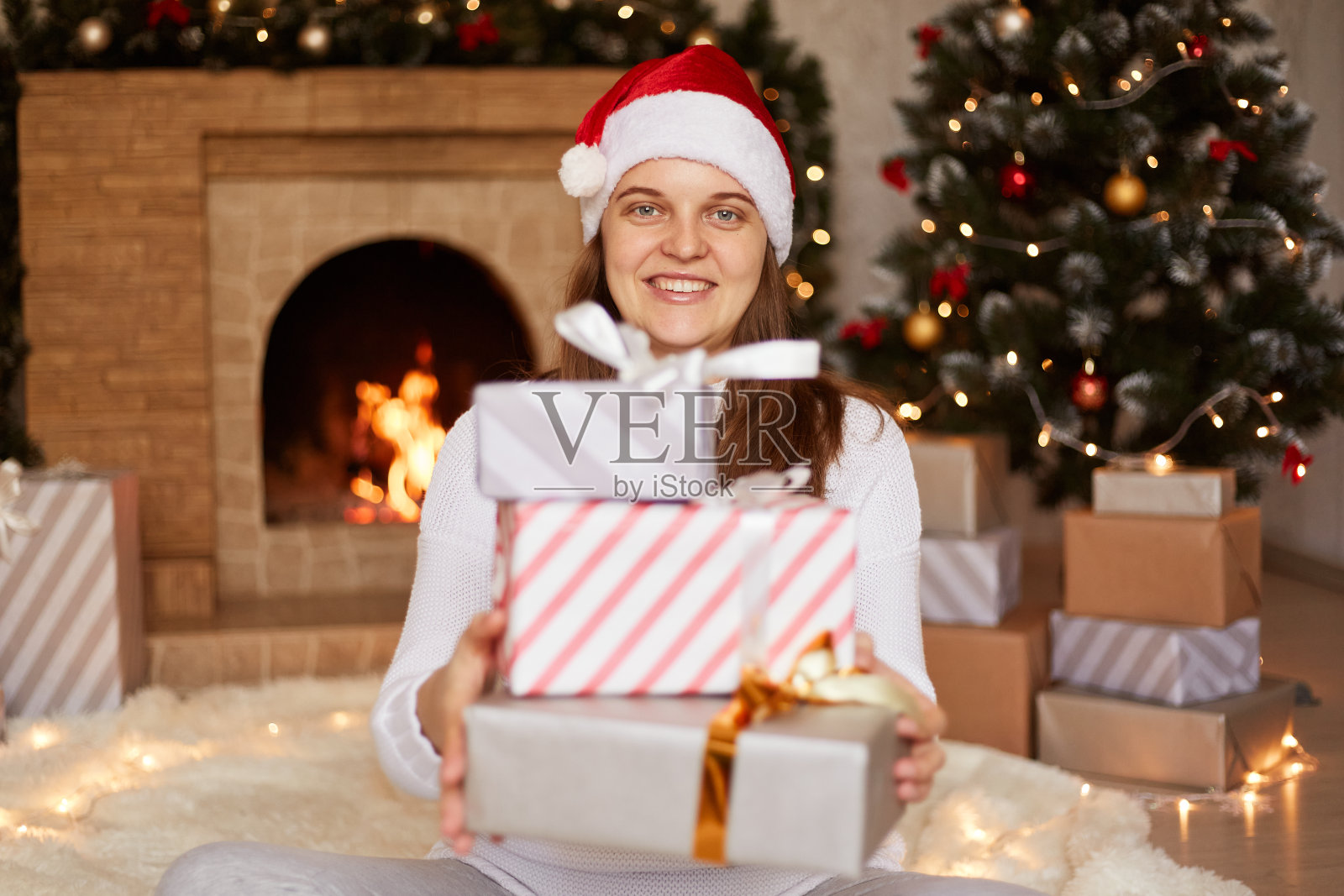戴着红色圣诞帽的快乐微笑的女人的肖像，拿着一堆礼物看着相机，庆祝圣诞礼物，在有壁炉的节日客厅摆姿势。照片摄影图片