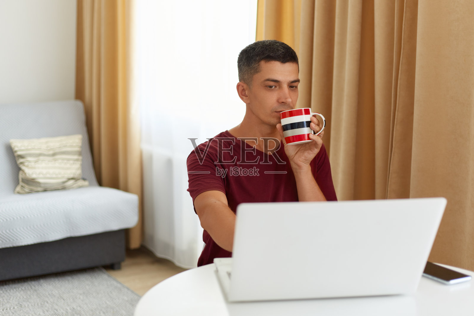冷静英俊的自由职业者穿着紫红色t恤坐在笔记本电脑前，享受着早上的热咖啡或茶，在沙发和窗帘的背景上摆姿势。照片摄影图片