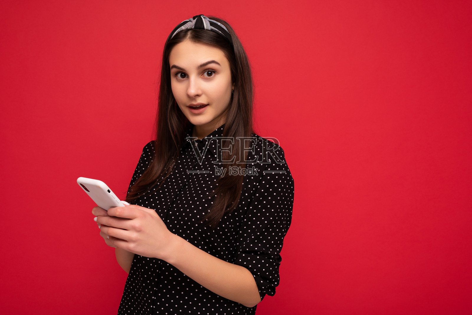 美丽的黑发女孩孤立地站在红色的墙上，穿着休闲时尚的黑色衣服，用手机写短信看着相机照片摄影图片