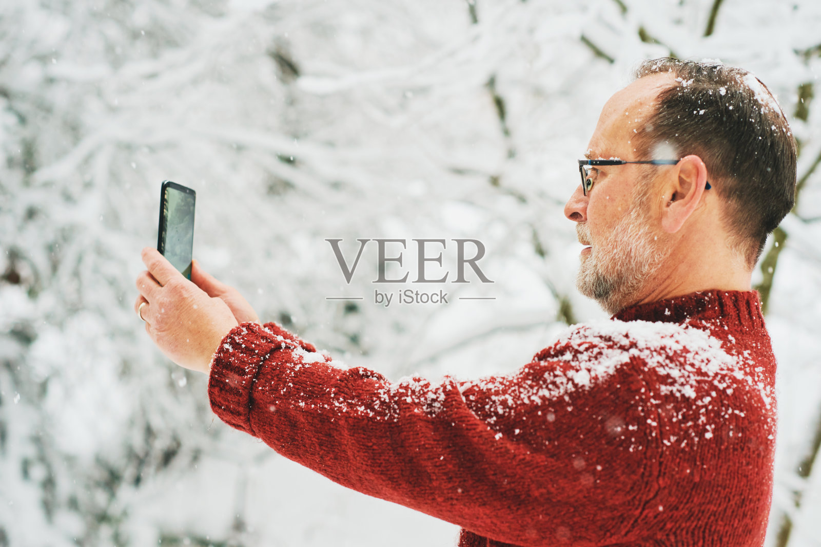 中年男子拿着智能手机，为社交媒体拍照或视频，冬天的森林下雪照片摄影图片