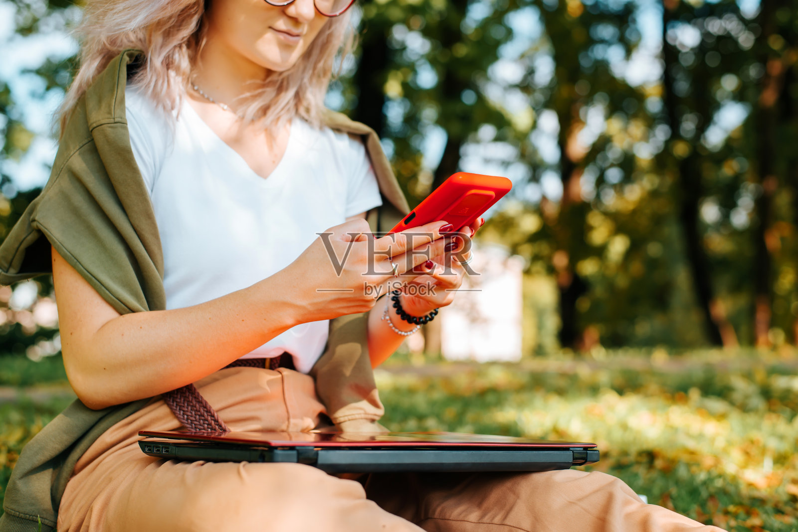 年轻的潮女戴着眼镜坐在公园里拿着手机和笔记本电脑，女学生在社交媒体上聊天，上网，在户外发信息。选择性关注智能手机照片摄影图片