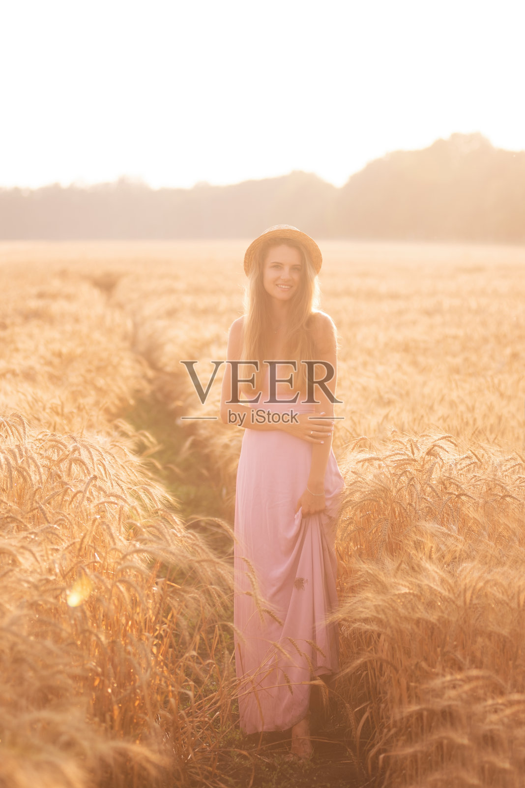 一个戴着草帽、穿着粉红色长裙的年轻快乐女人的肖像站在金色的麦田里照片摄影图片