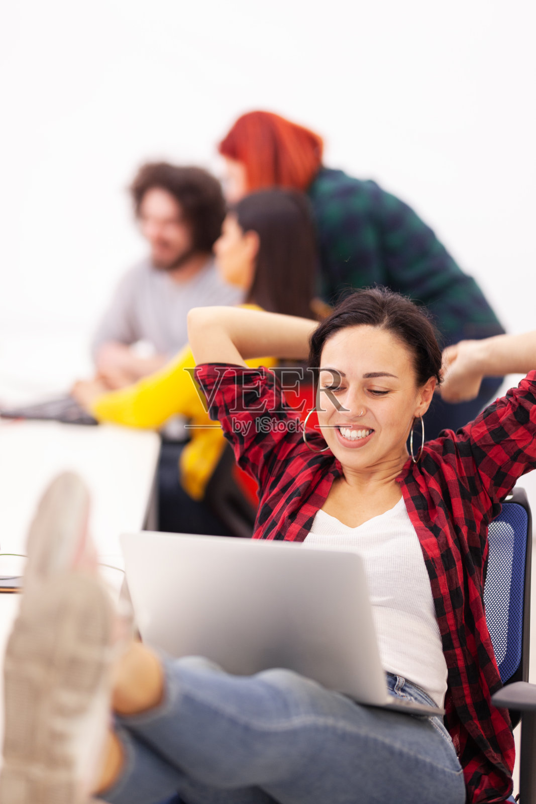 在一个现代的共享办公空间里，漂亮的女孩在桌子上用笔记本电脑工作。快乐的成功的手势。后面是业务上的同事照片摄影图片