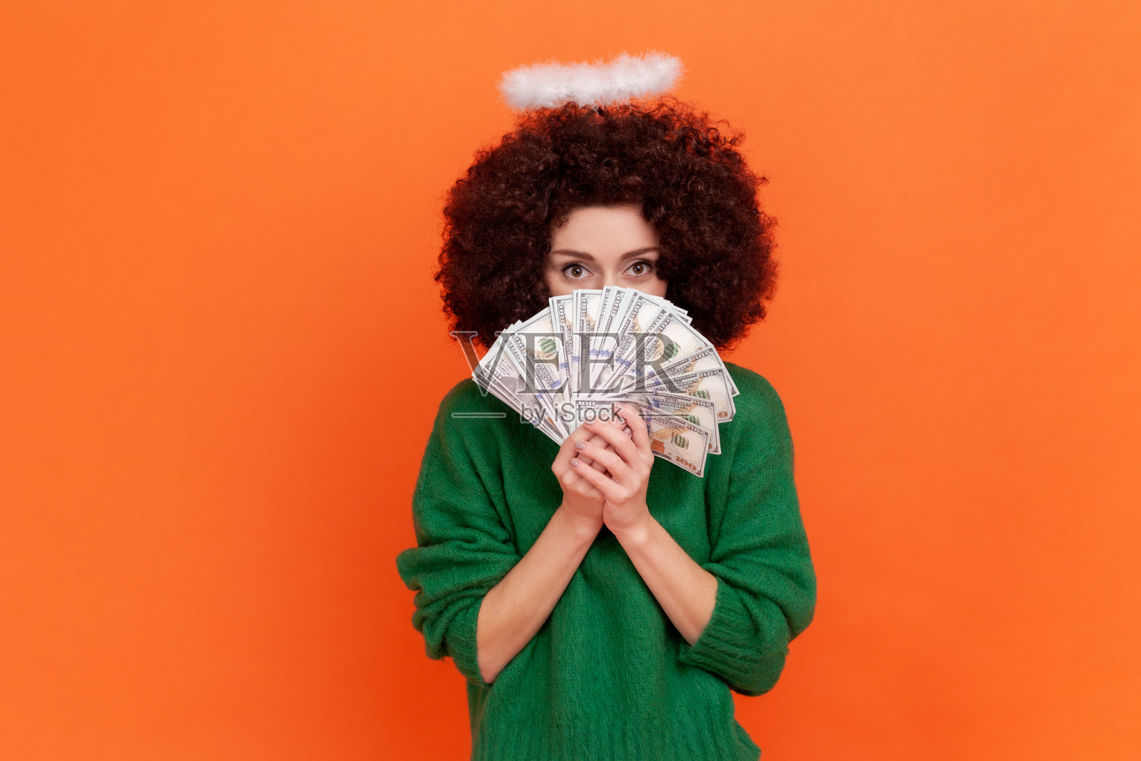 天使般的女人的肖像，留着非洲式发型，穿着绿色休闲风格的毛衣，用大扇子的美元钞票盖住半张脸。照片摄影图片