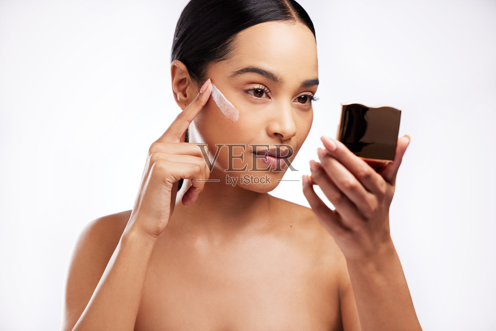 摄影室拍摄了一个美丽的年轻女子在白色背景下用镜子给她的脸涂润肤霜照片摄影图片