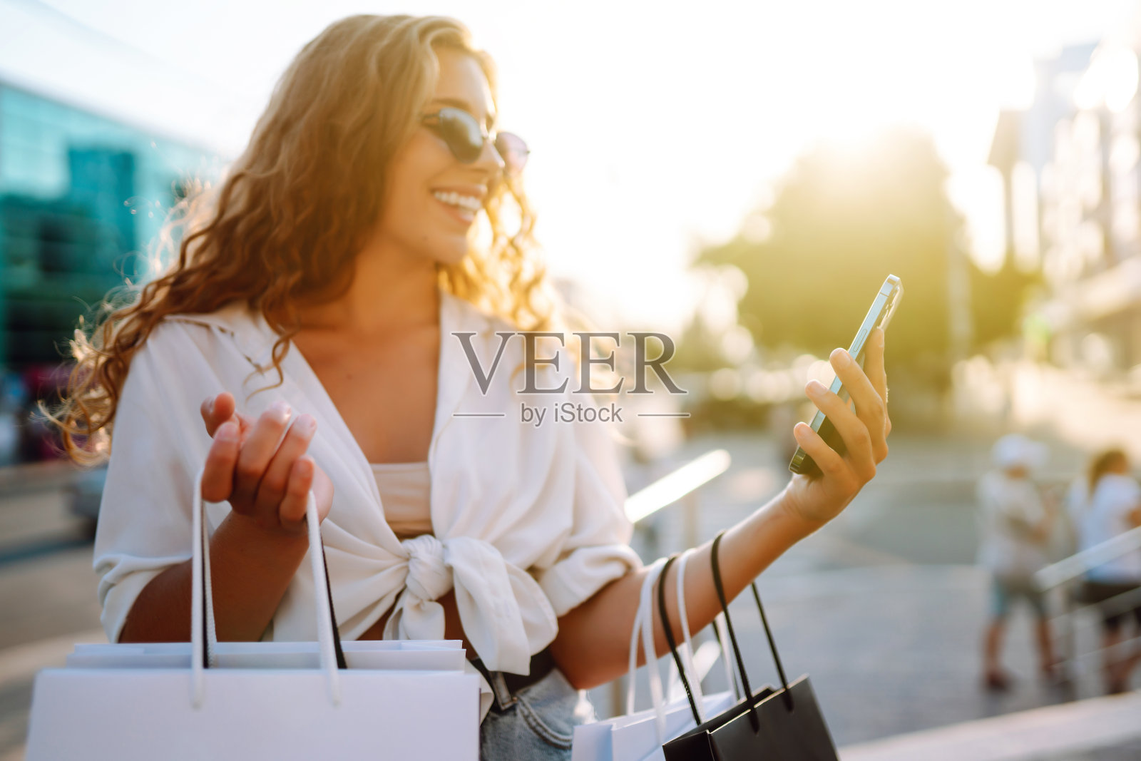 年轻女子用手机，拿着购物袋，站在户外，挑选和订购衣服和礼物。​照片摄影图片