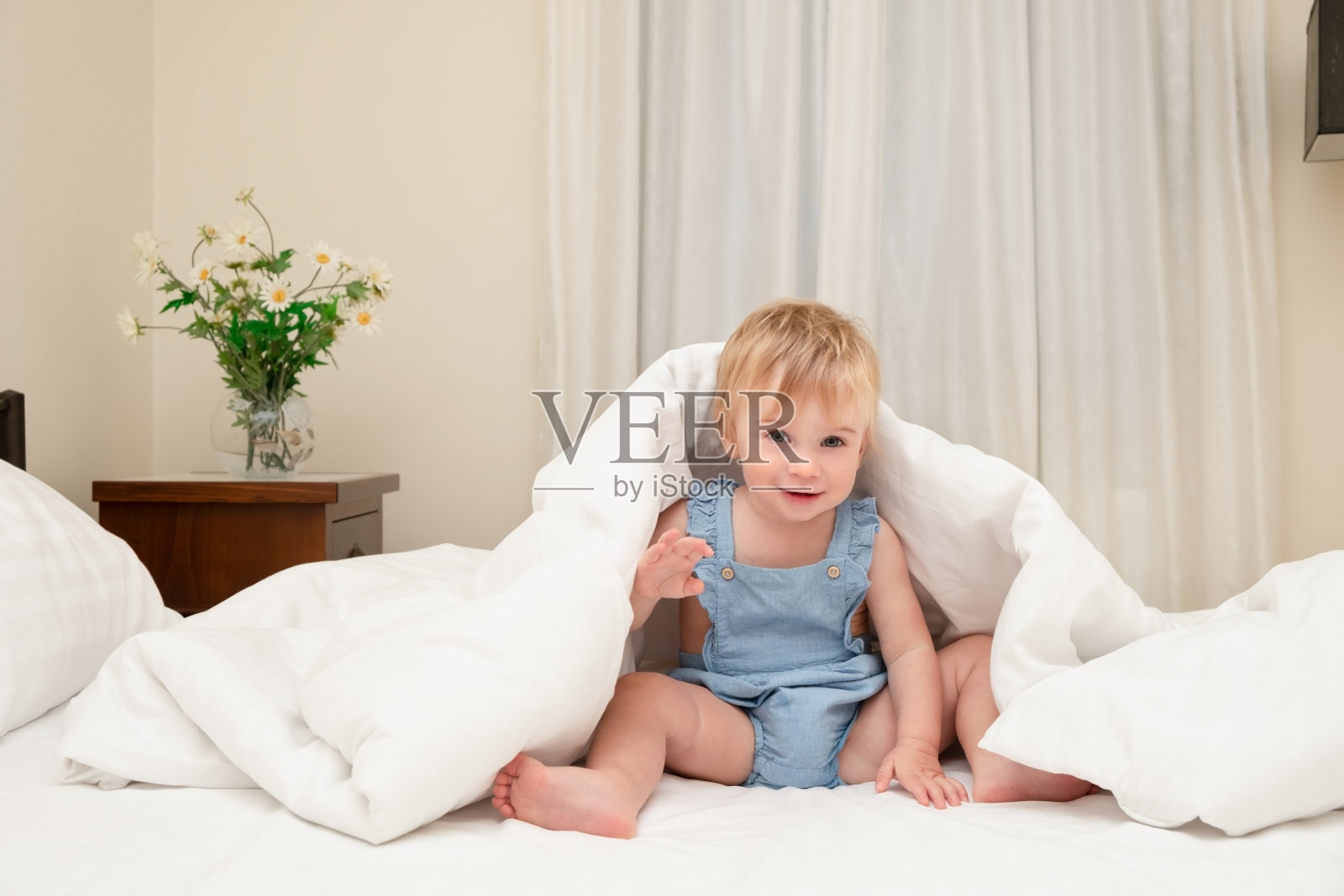 可爱的白种人金发小女孩玩耍，在床上温暖的毯子下的白色床单上玩耍。1-2岁的婴儿微笑着看着相机。可爱的寒冷孩子的概念在秋天，冬天照片摄影图片