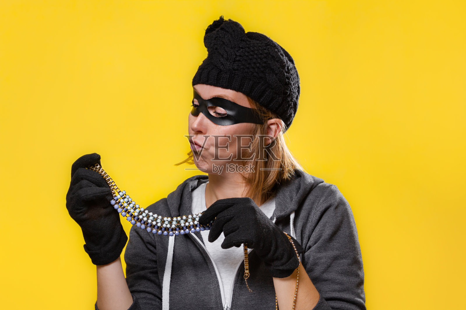 盗窃的概念。一个戴着帽子、面具和手套的女人双手捧着珠宝，仔细端详着。黄色背景和复制空间照片摄影图片