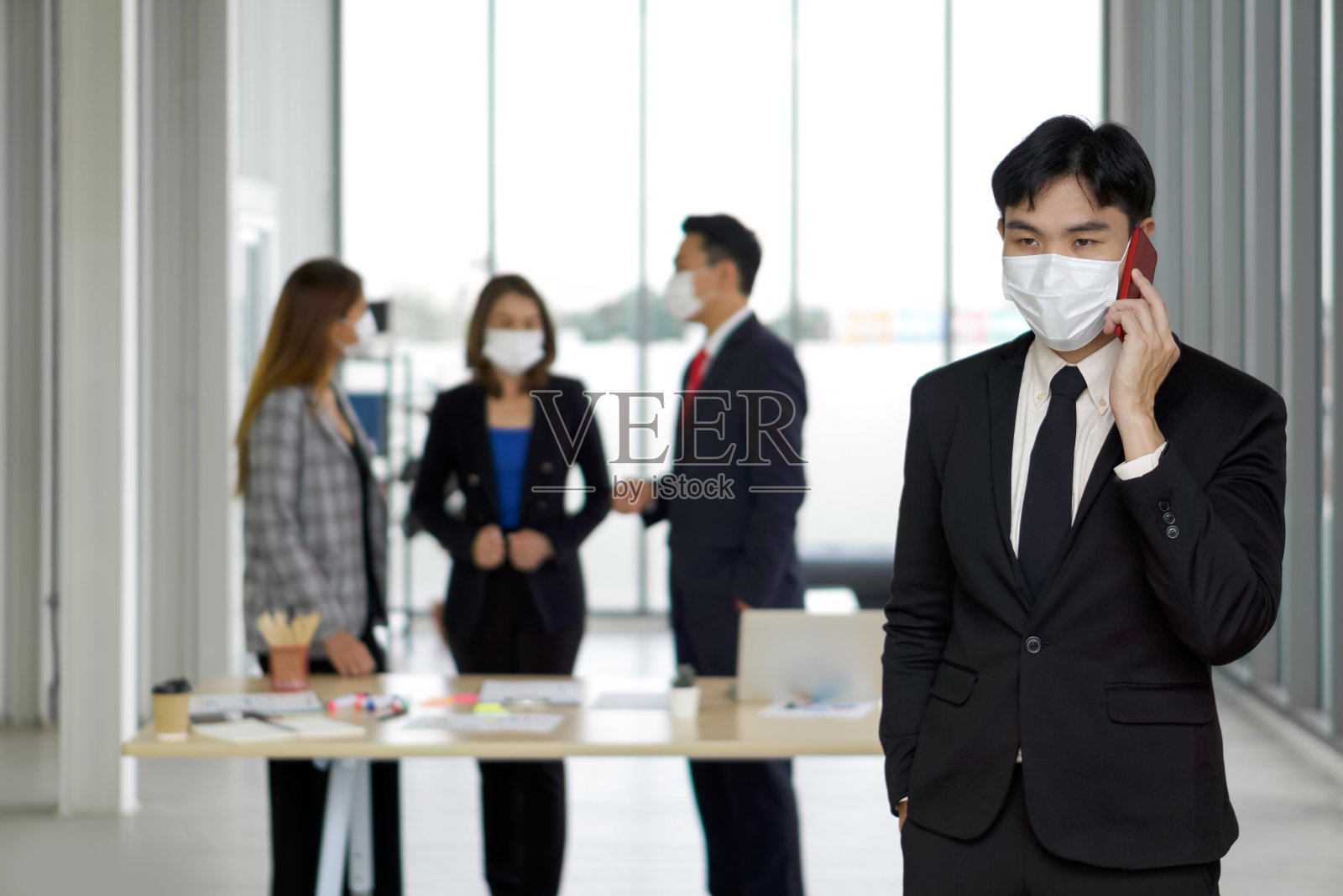 年轻的亚洲商人通过移动电话与商业伙伴交谈。团队正在后台为会议做准备。每个人都戴着口罩，防止办公室里的冠状病毒爆发。照片摄影图片