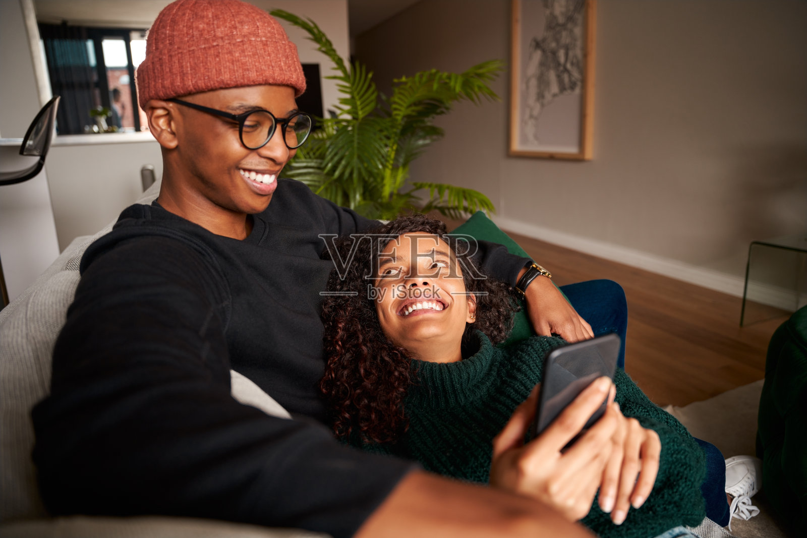 时尚的多民族情侣边看手机边微笑。沙发上的现代公寓照片摄影图片