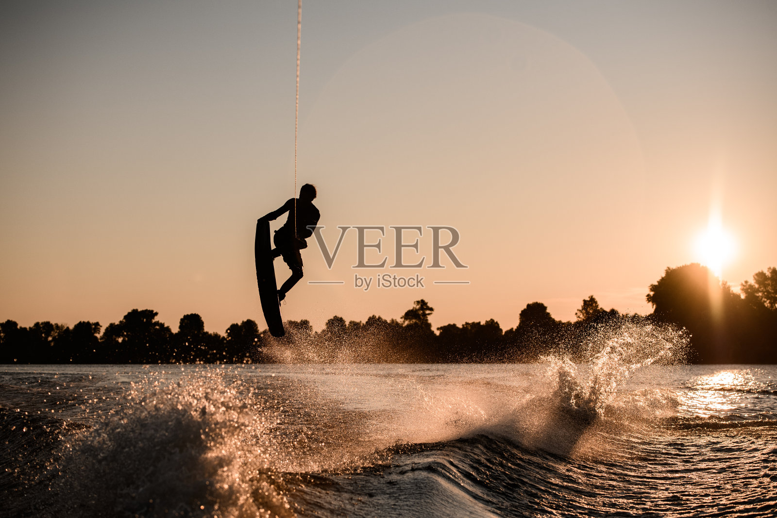 日落时分，活跃的男骑手手持绳索，在水上滑水板上进行极限跳跃的黑色剪影。照片摄影图片