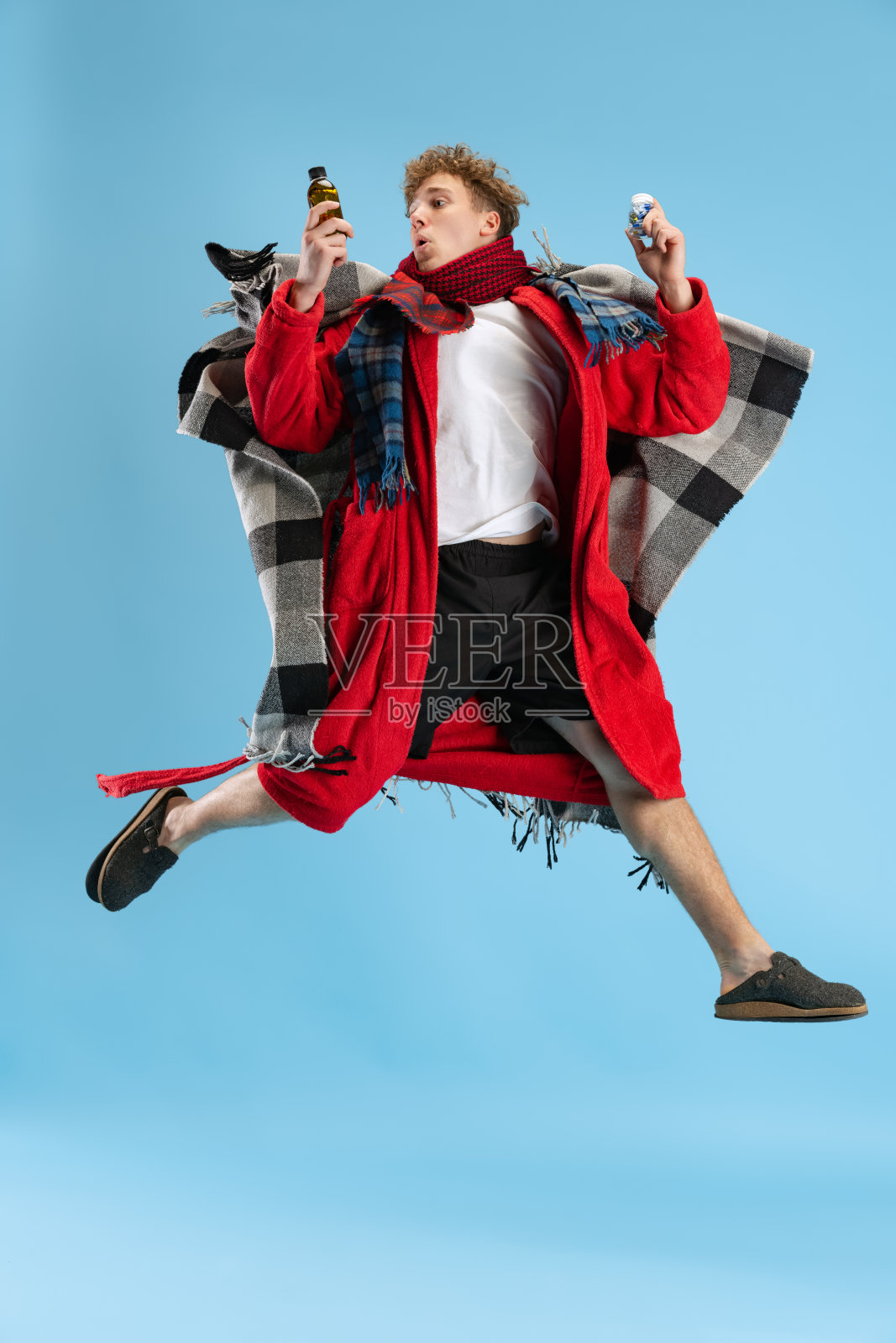 生病的小男孩的全身肖像，穿着格子呢，长袍和围巾快乐地跳过蓝色背景。感冒，流感，过敏照片摄影图片