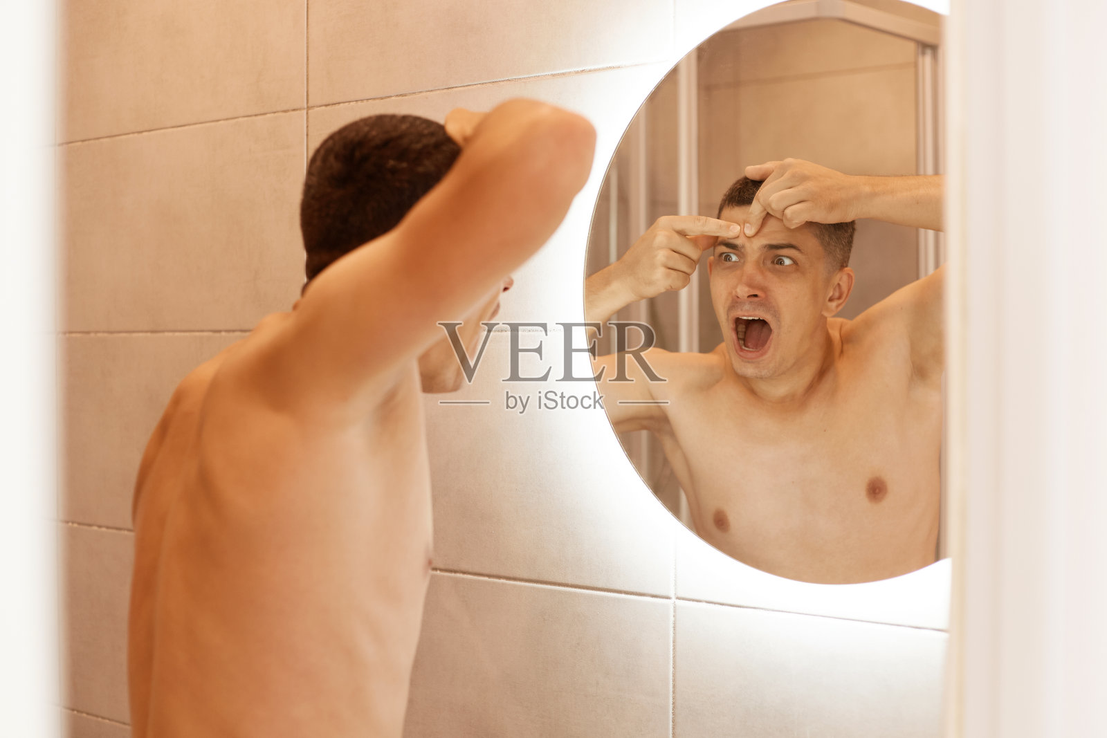 在室内拍摄的照片中，一个年轻的黑发男子对着镜子尖叫，因为它发现了皮肤上的问题，发现了额头上的痤疮，嘴巴张得大大的。照片摄影图片