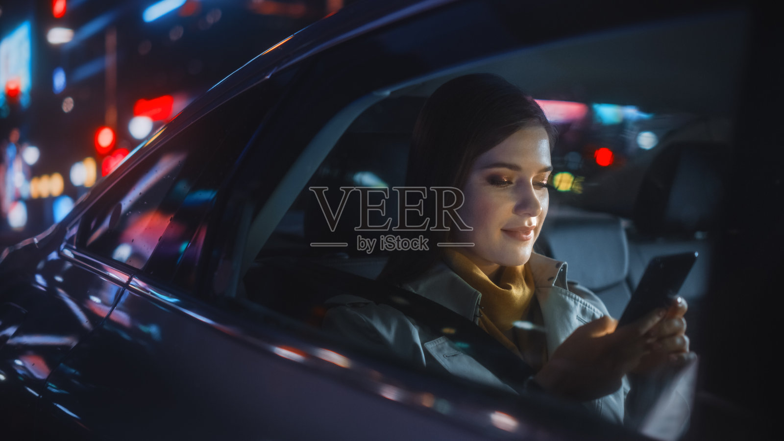 时尚女性在晚上乘出租车回家。美丽的女乘客在城市街道上使用智能手机，霓虹灯还在工作。照片摄影图片