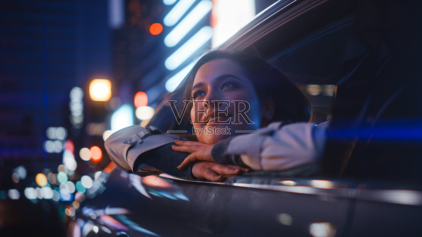 兴奋的年轻女性坐在后座上，晚上回家。看着窗外，惊叹城市街道的霓虹灯招牌是多么的美丽。照片摄影图片