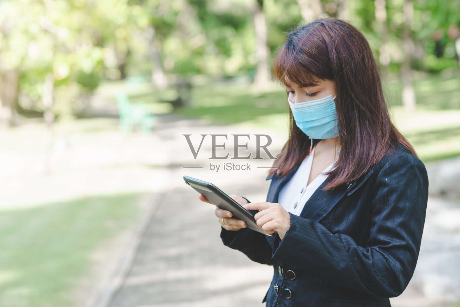 商业女性戴着口罩，用智能手机平板电脑在屏幕上打字，在有日出和模糊自然背景的公园里使用应用程序设备。由于冠状病毒或covid-19，商业进入新常态。照片摄影图片