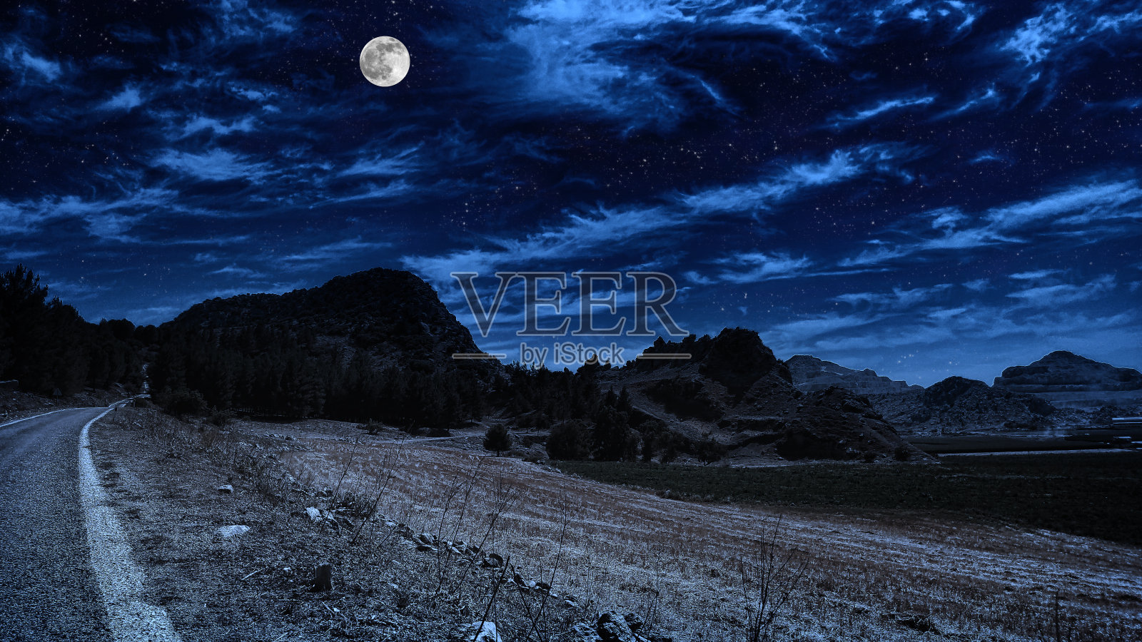 古老的城堡在山上的夜晚。山脊的岩石山峰在遥远的背景中，在满月的光和多云的天空。照片摄影图片