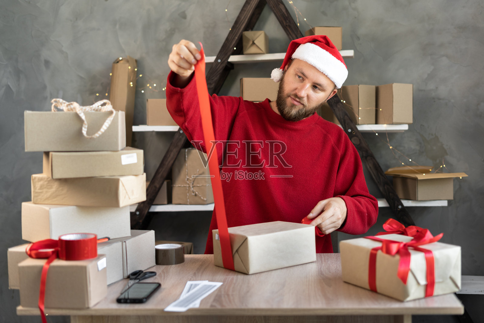 一名戴着圣诞帽的男子在仓库里准备圣诞礼物并系上红丝带。桌子上有纸板箱。新的一年销售照片摄影图片