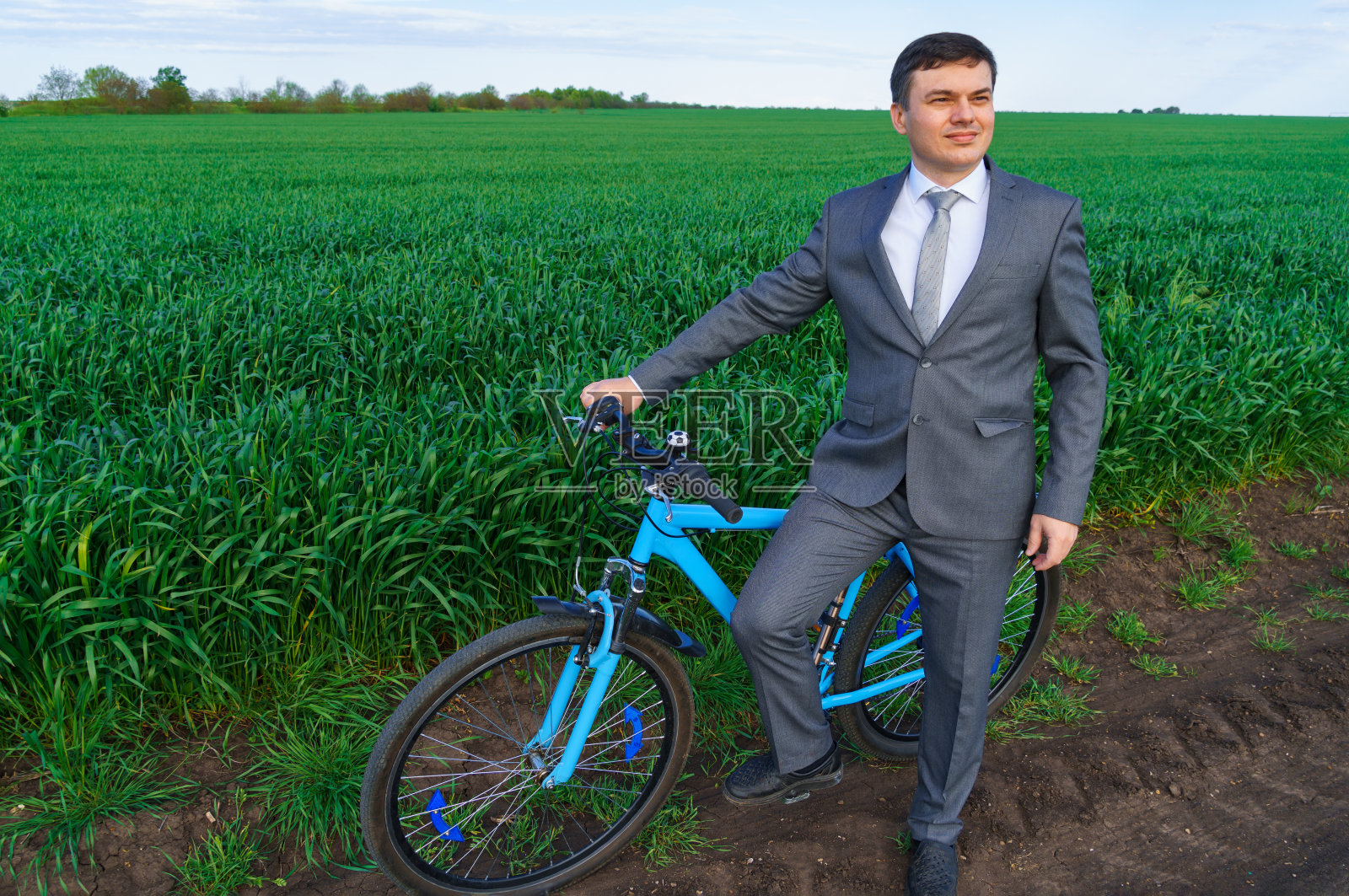 一个商人骑着自行车在麦田里摆姿势，绿色的草地和蓝色的天空为背景照片摄影图片