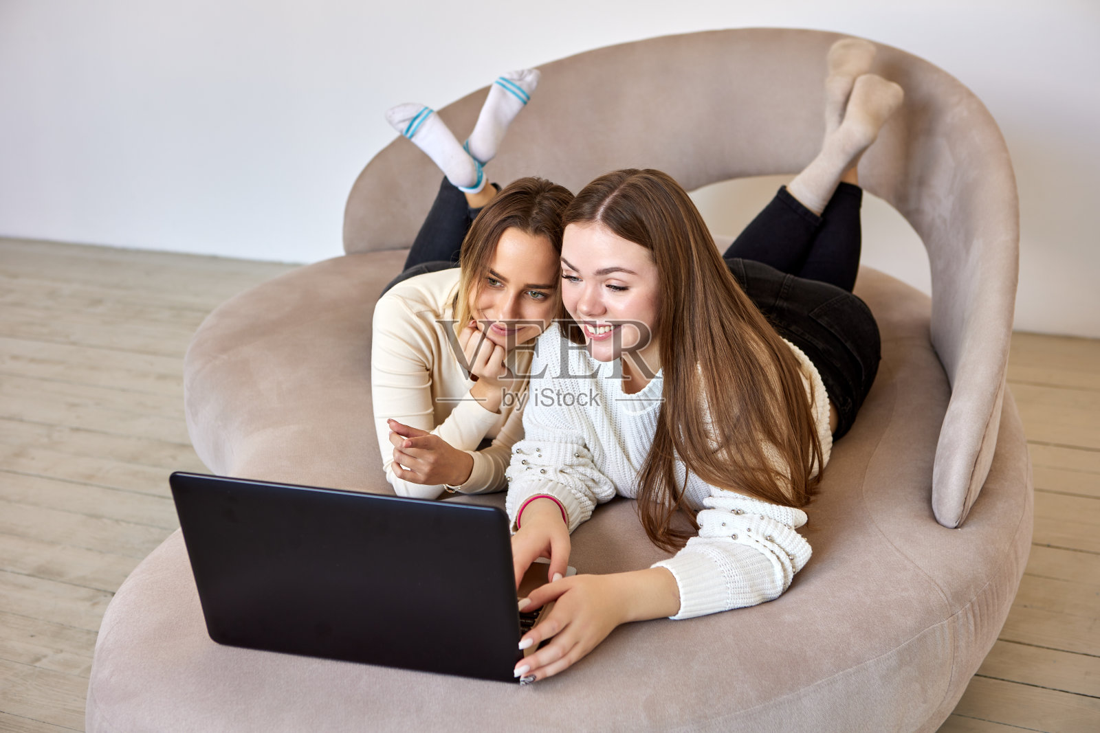 两个面带微笑的姐妹躺在沙发上，拿着笔记本电脑远程工作。照片摄影图片
