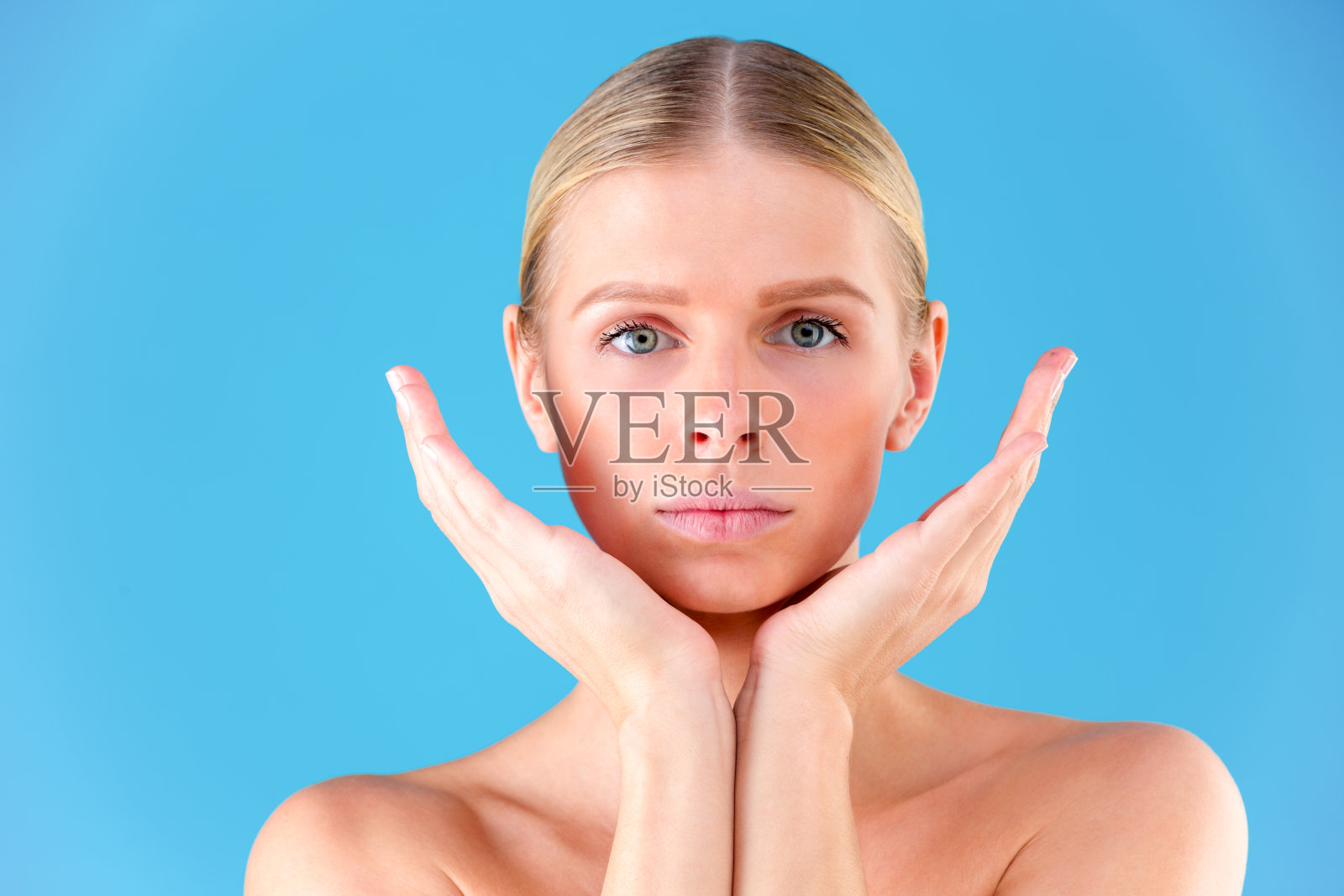 接近的肖像华丽的美丽和好看的年轻女人与无瑕的皮肤。她用手捂住孤立在蓝色背景上的脸颊照片摄影图片