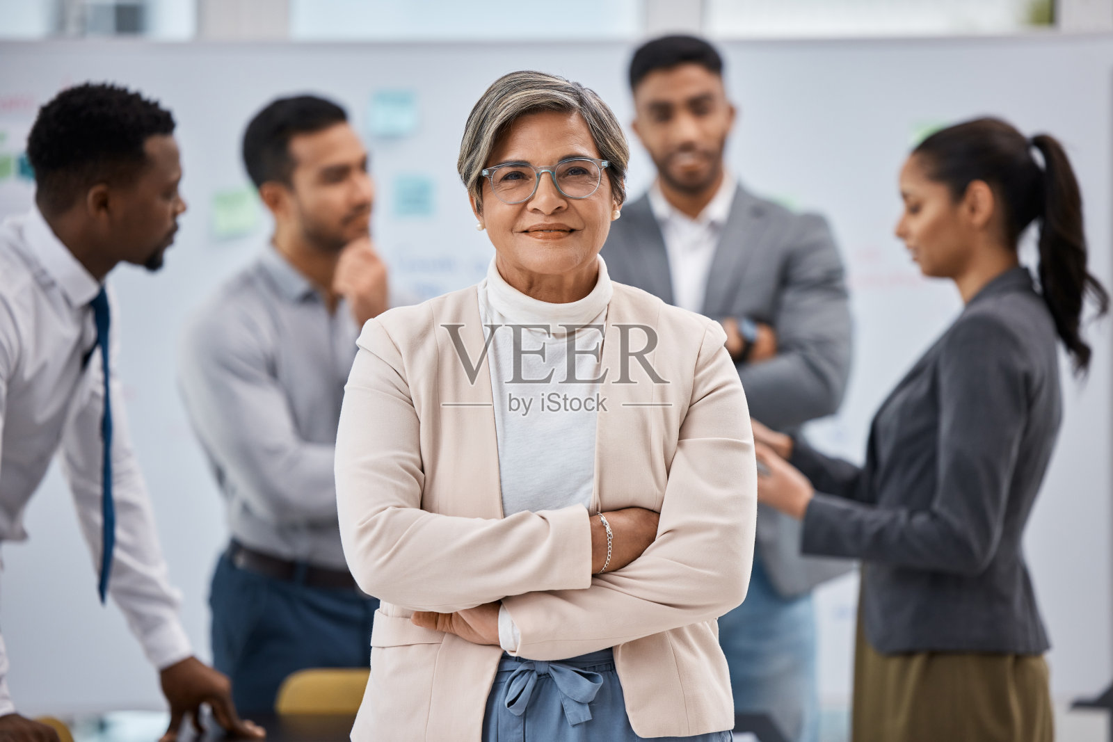 一位成熟的女商人双臂交叉站在办公室里，身后是她的同事照片摄影图片
