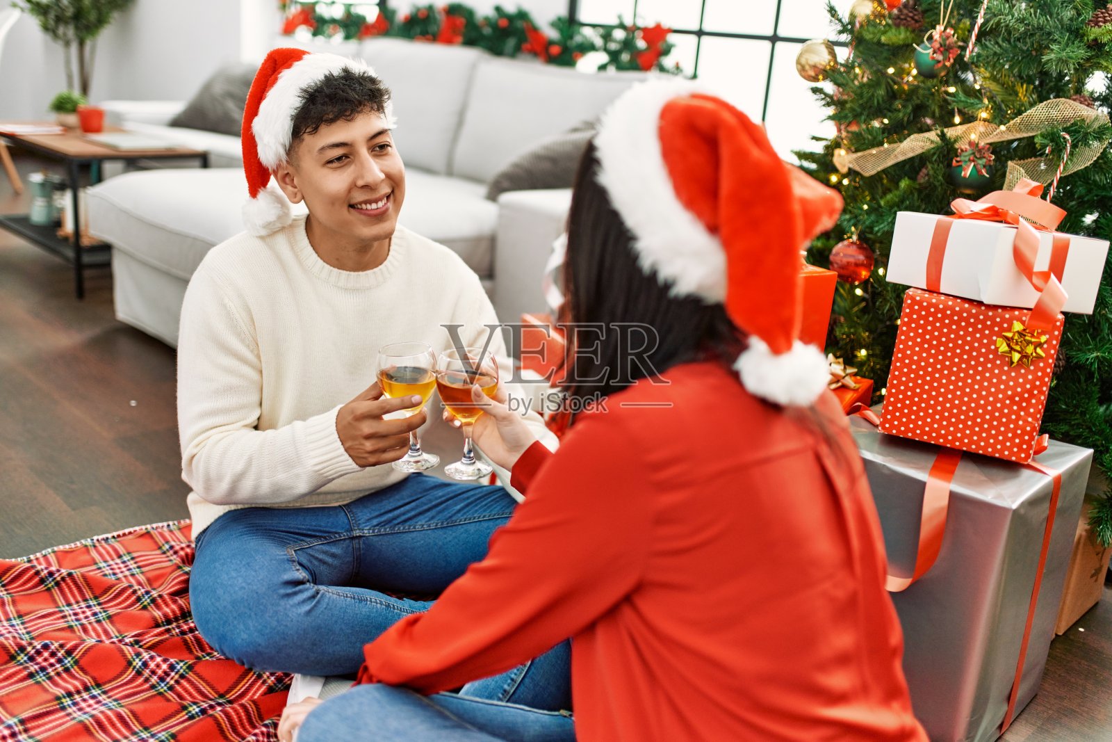 年轻的西班牙夫妇微笑幸福戴着圣诞帽在家里用香槟祝酒。照片摄影图片