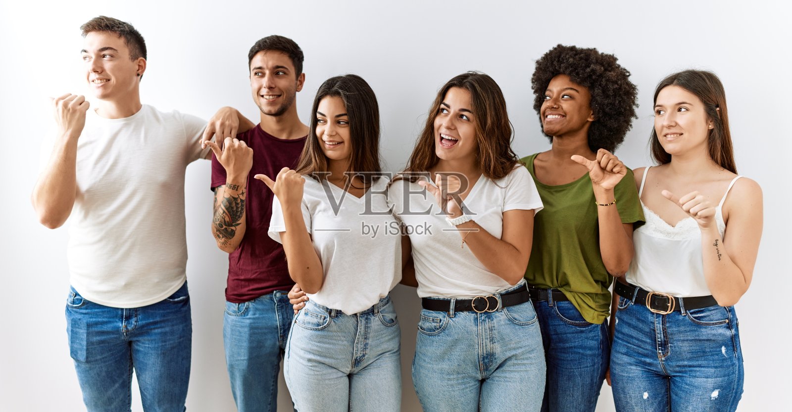 一群年轻的朋友站在一个孤立的背景下，笑脸相迎，拇指向上指着一边。照片摄影图片