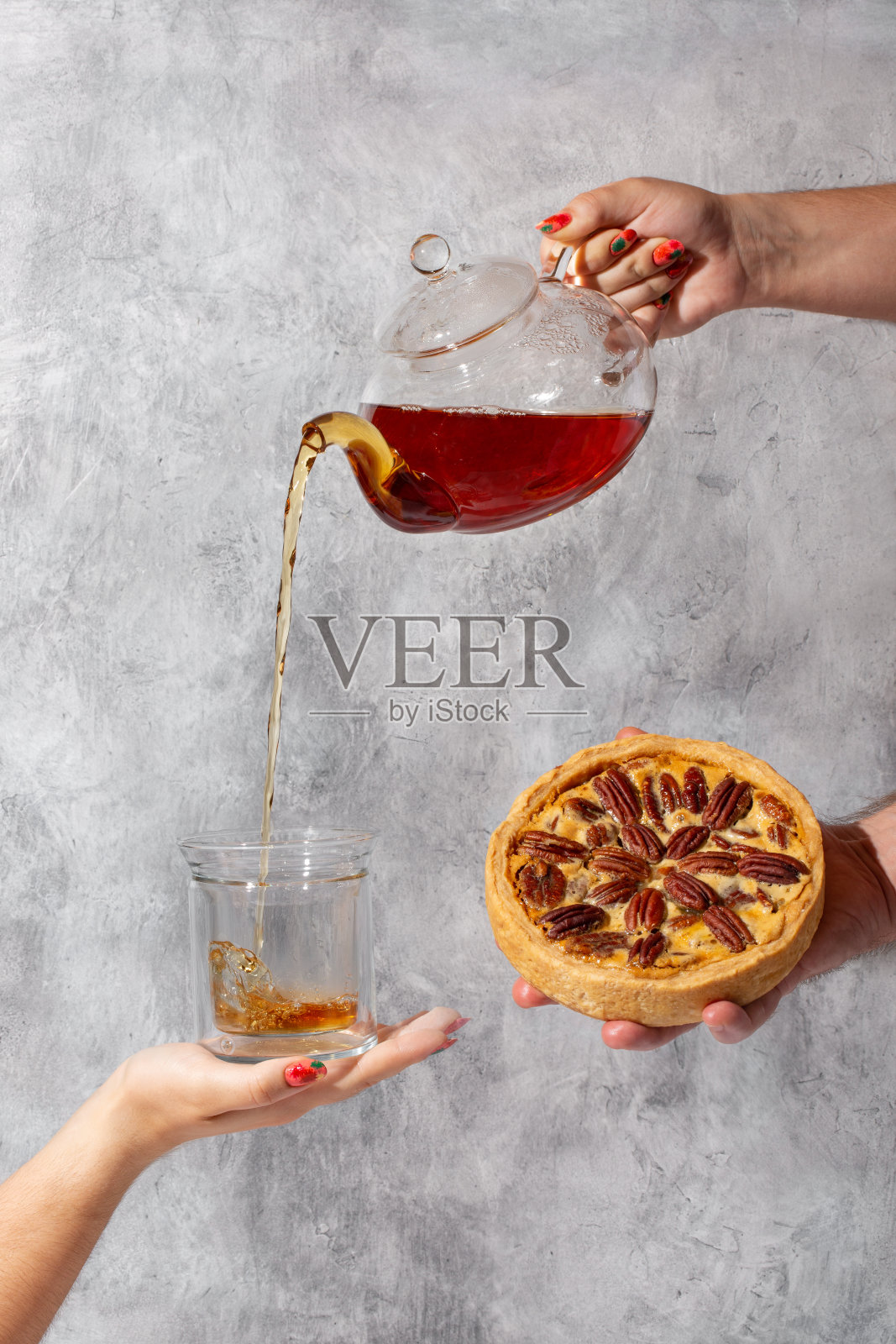 核桃派茶话会。倒茶的创意镜头。女手倒茶从玻璃茶壶到杯子在混凝土灰色背景。照片摄影图片