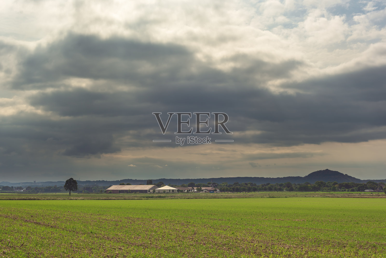 在多云的天空下，地平线上刚刚播种的、有谷仓和树木的田地。照片摄影图片