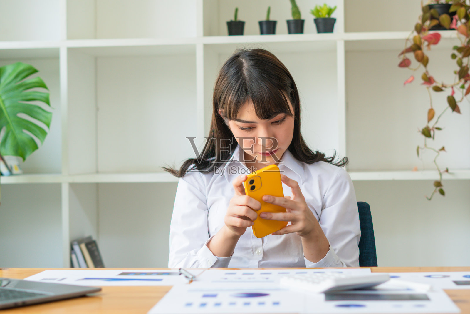 快乐的年轻亚洲女人讲手机和微笑，而坐在她的工作地点在办公室。照片摄影图片