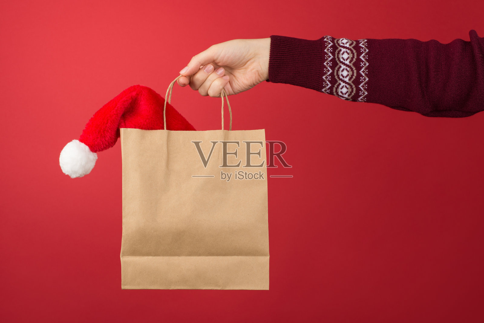短特写照片的女性手在红色和白色的圣诞毛衣持有工艺纸袋与圣诞老人的帽子在孤立的红色背景与copyspace照片摄影图片