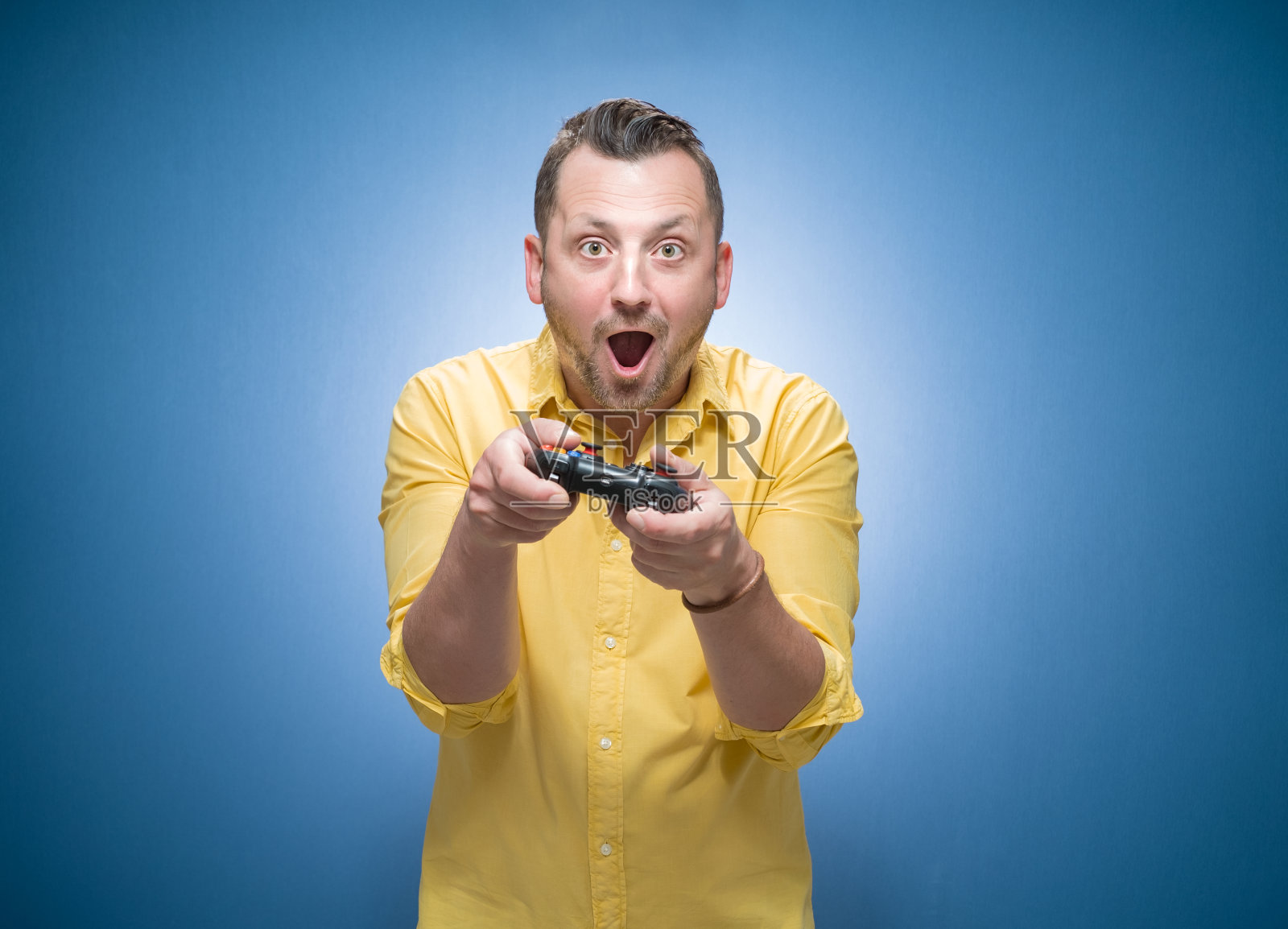一名身穿黄色衬衫的男子在蓝色背景下拿着操纵杆玩电子游戏。惊讶的家伙拿着游戏控制器看着摄像机照片摄影图片