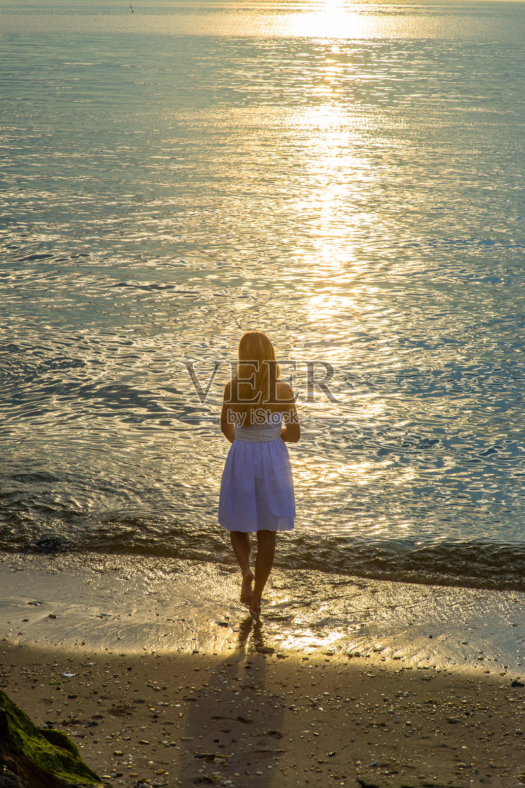 年轻女子站在沙滩上的水里，看着日落照片摄影图片
