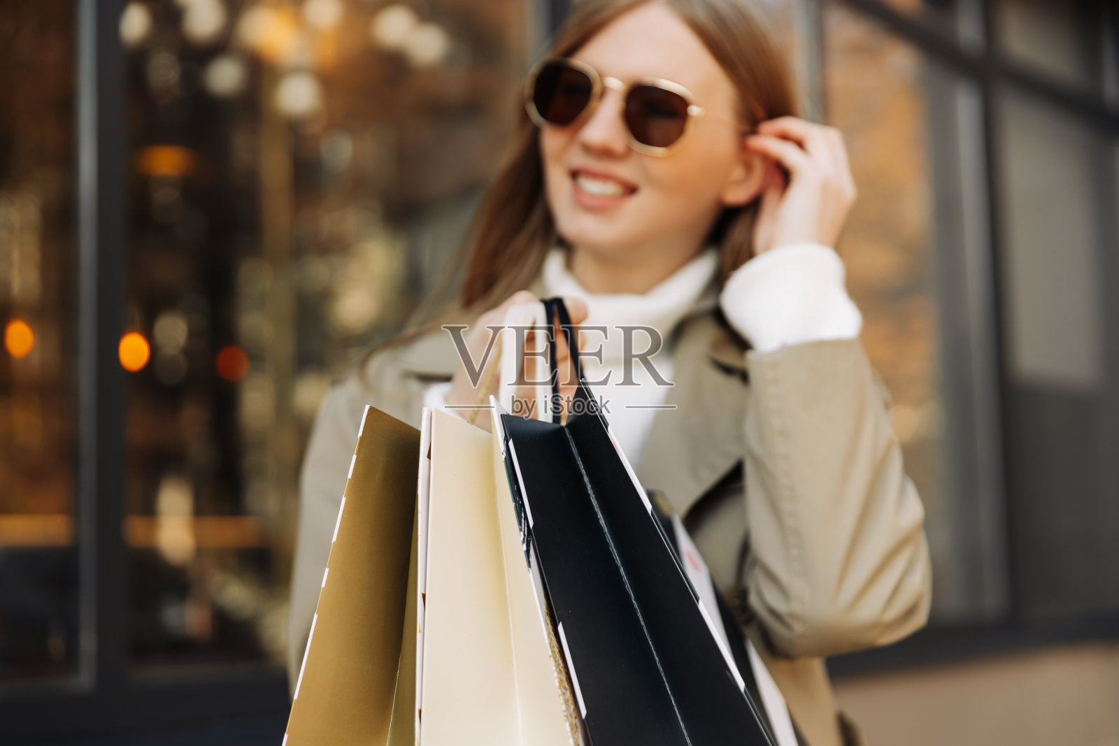 美丽的女人戴着太阳眼镜，穿着米色风雨衣，手里拿着购物袋，站在前面照片摄影图片