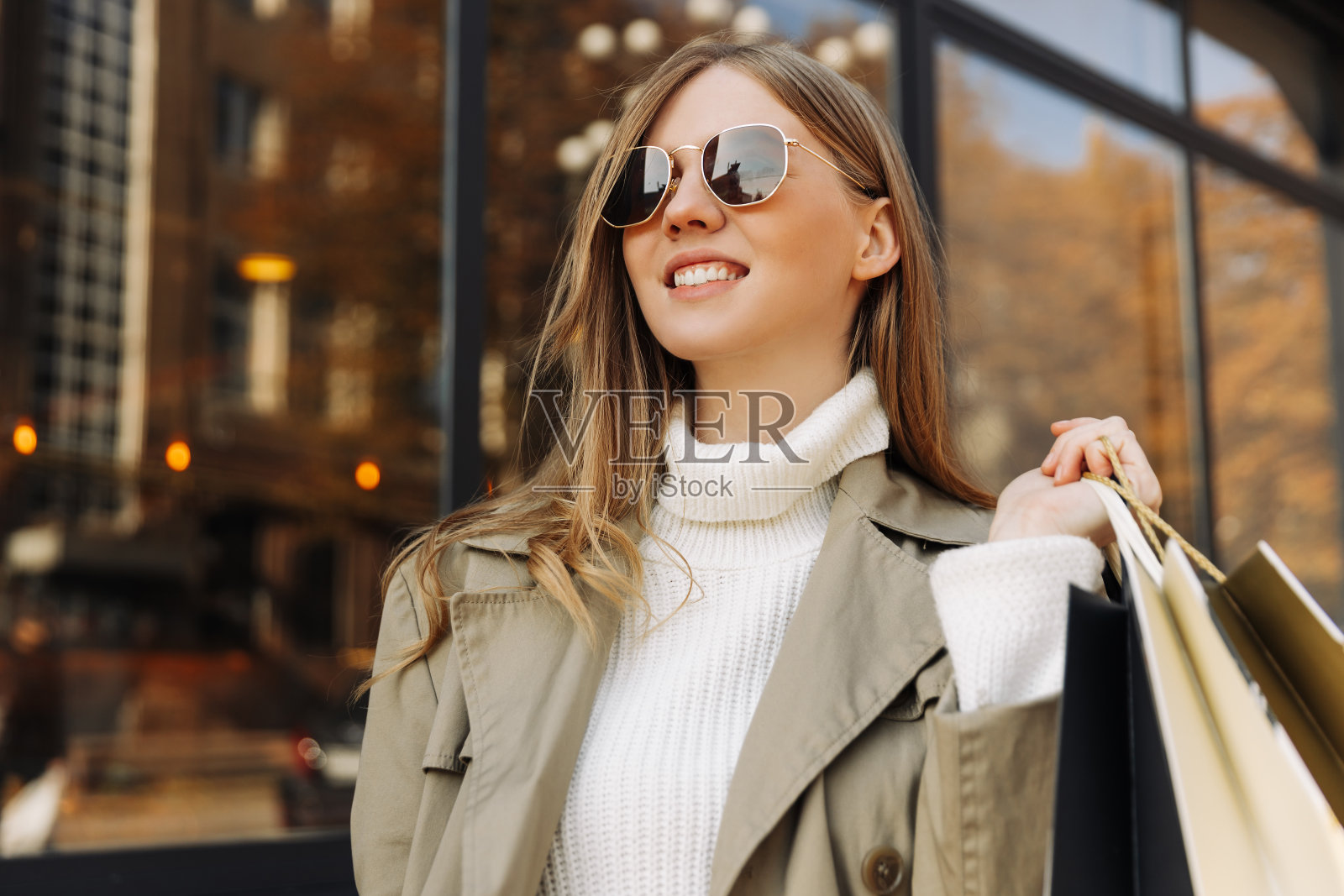 美丽的女人戴着太阳眼镜，穿着米色风雨衣，手里拿着购物袋，站在前面照片摄影图片