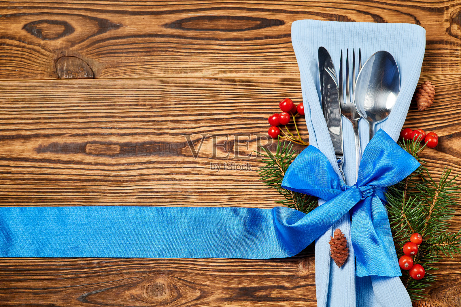 餐刀、叉子和勺子放在餐巾上，蓝色丝带系在木制背景上，木制背景上装饰着冷杉树枝、冷杉球果和花楸果照片摄影图片