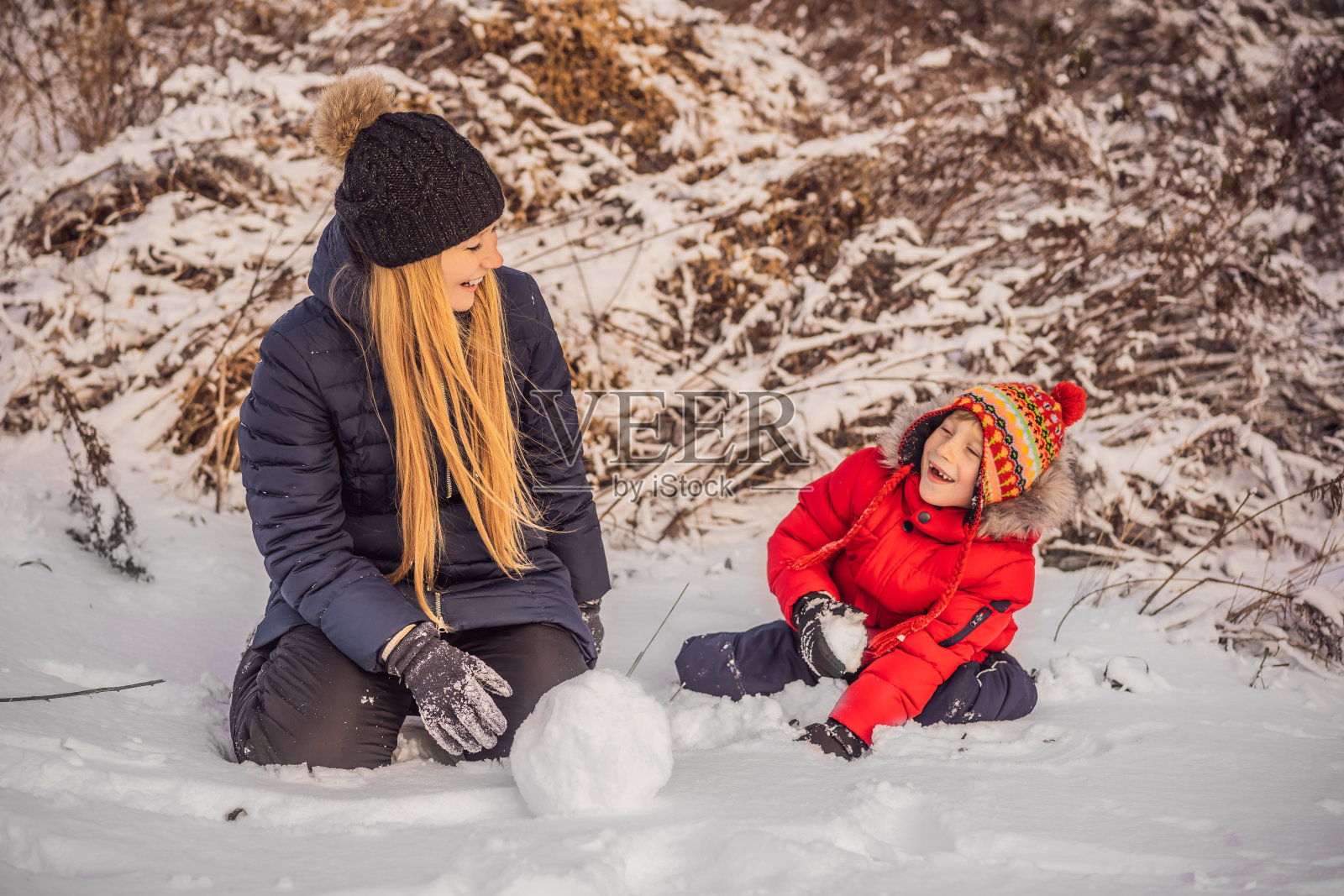 幸福的家庭穿着温暖的衣服。微笑的母亲和儿子在户外堆雪人。冬季活动的概念照片摄影图片