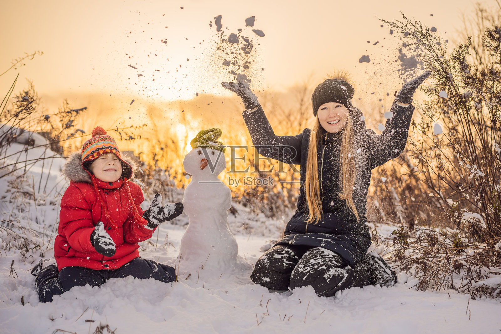 幸福的家庭穿着温暖的衣服。微笑的母亲和儿子在户外堆雪人。冬季活动的概念照片摄影图片