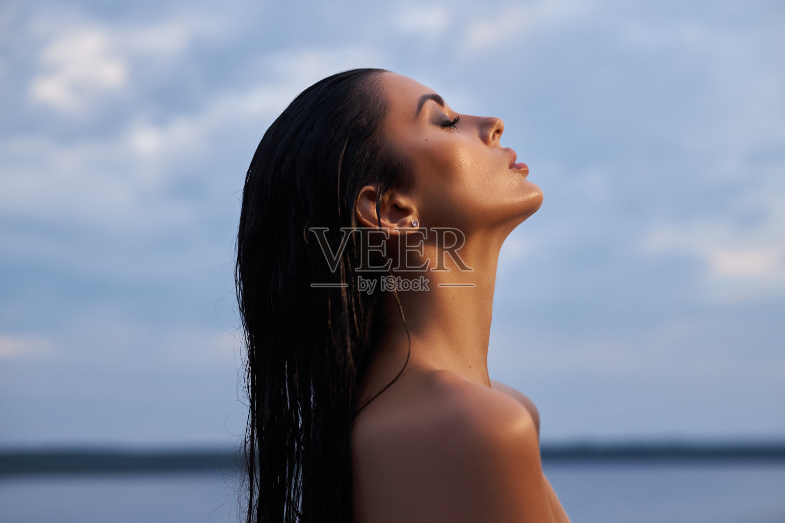 美丽的黑发女子，身穿黑色连衣裙，在蓝天背景下的海湖上摆姿势。长发性感的女人和美丽美丽的化妆在她的脸上照片摄影图片
