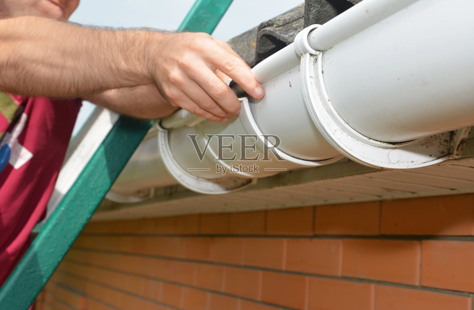 天沟安装。一名男子正在梯子上安装屋顶排水沟u2019滑动接头，以连接排水沟，防止漏水。照片摄影图片